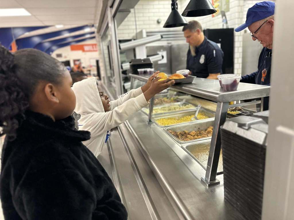 Two children are standing in line in a school cafeteria being served food by two district staff members. One of the children is reaching for a tray of food that has corn and chicken on it.