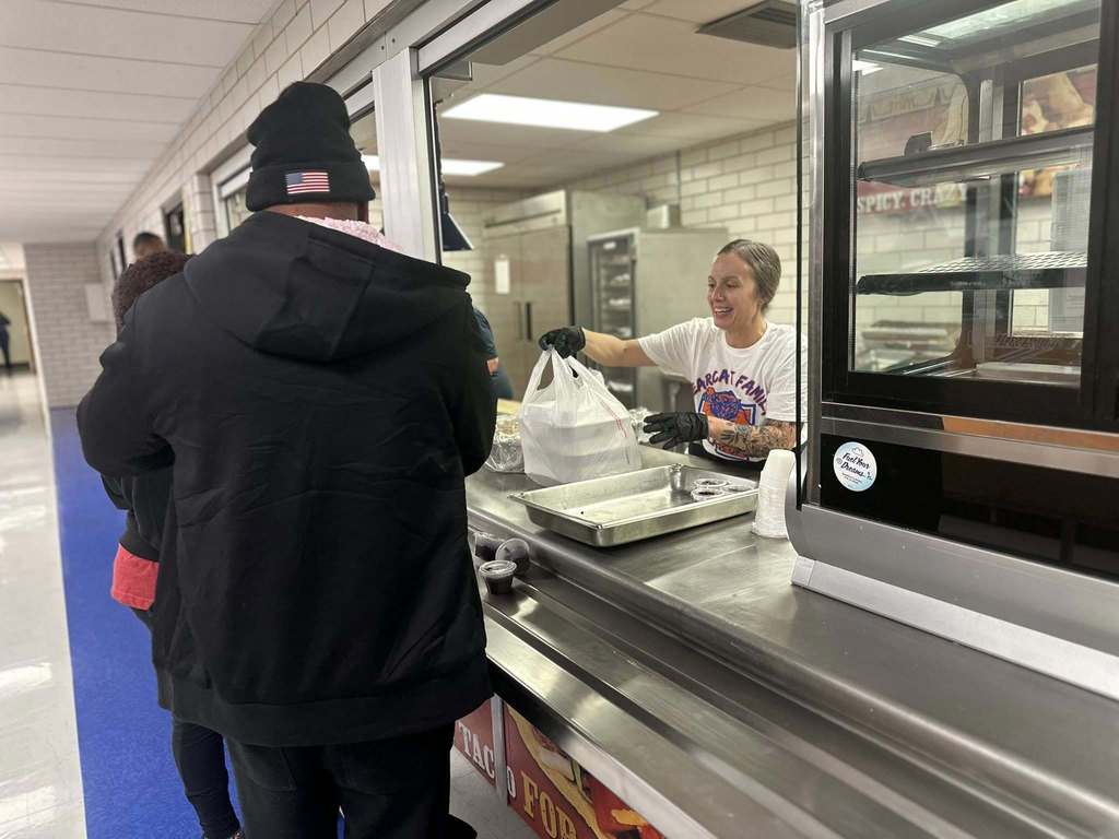 A district staff member is handing a bagged meal through a serving window to a man. 