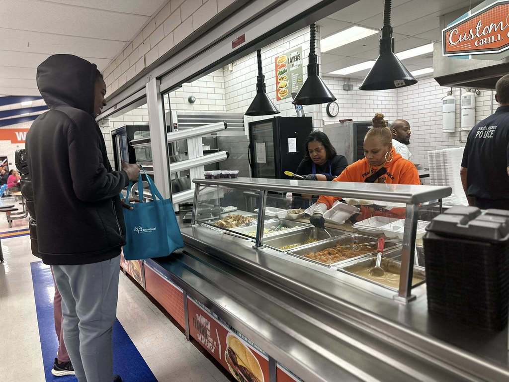 A man in a hoodie is standing in line in a school cafeteria, holding a blue tote bag. Dr. Andrea Berry-Brown, Superintendent of Schools and a district staff member are serving food behind a glass counter. Other people can be seen in the background.