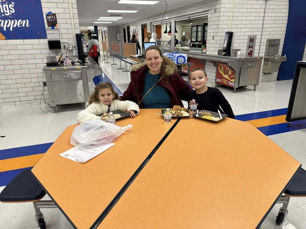 A family of three, including an adult woman and two children, are sitting at a table in a school cafeteria enjoying a meal together.
