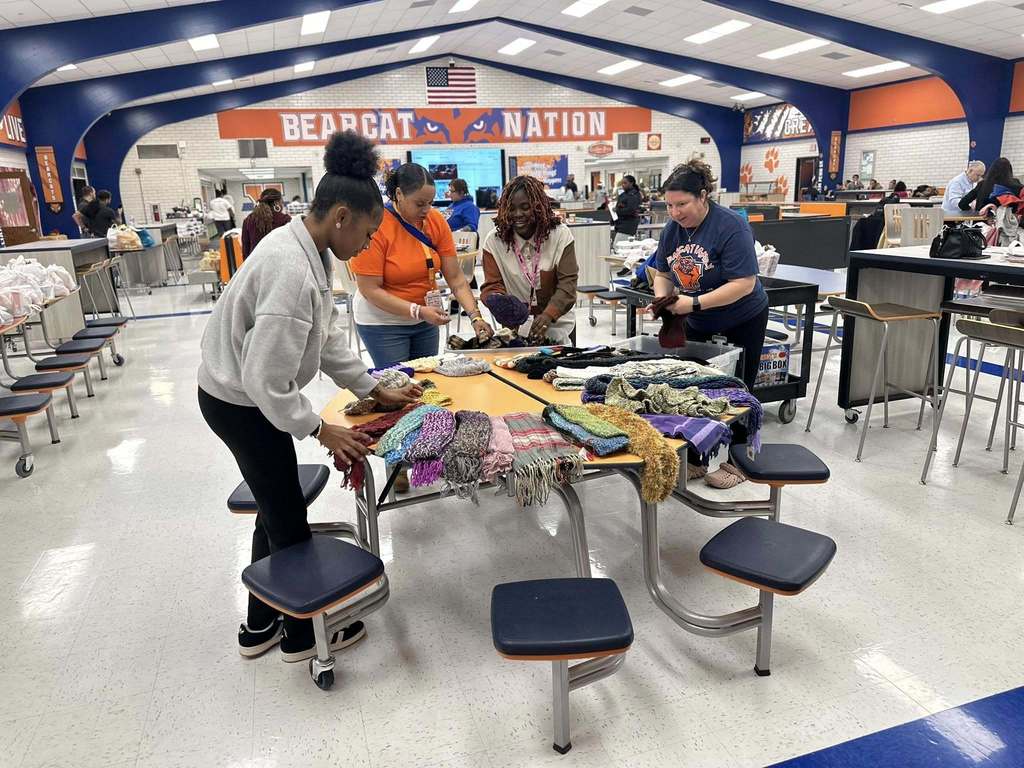 A group of four district staff members are organizing knitted scarves on a table in a school cafeteria. Behind them is a sign that reads "BEARCAT NATION" on the wall.
