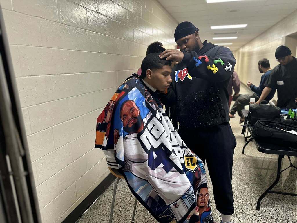 A barber is trimming a young man's hair in a school hallway, with other people in the background.