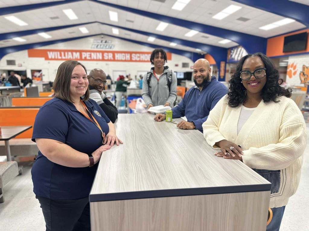 A group of district staff members and a teenage boy are standing and sitting at a table in a school cafeteria. Behind them is a sign that reads "William Penn Sr. High School" on the wall.