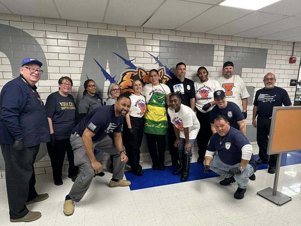 A group of district staff members, including York City School Police are standing together in front of a mural of a roaring bear head in a school cafeteria.