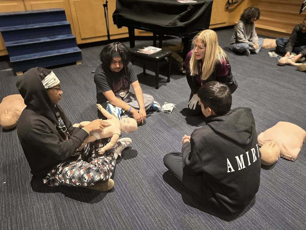 A group of three students and one adult are sitting on a dark carpeted floor in a school auditorium. One of the students is holding an infant mannequin, practicing CPR. Other people can be seen in the background, also performing CPR. 