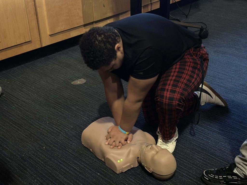 A student is keeling down on a dark carpeted floor in a school auditorium, practicing CPR on a mannequin.