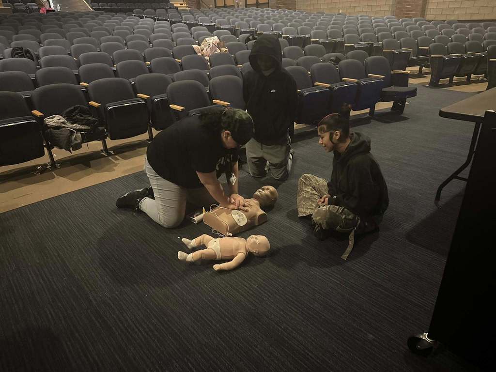 Three students are kneeling down on a dark carpeted floor in a school auditorium, practicing CPR on a mannequin. An infant mannequin can be seen nearby. 