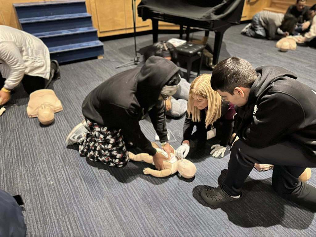 A group of four students and one adult are sitting down on a dark carpeted floor in a school auditorium, practicing CPR on an infant mannequin. Other people can be seen in the background, also practicing CPR.