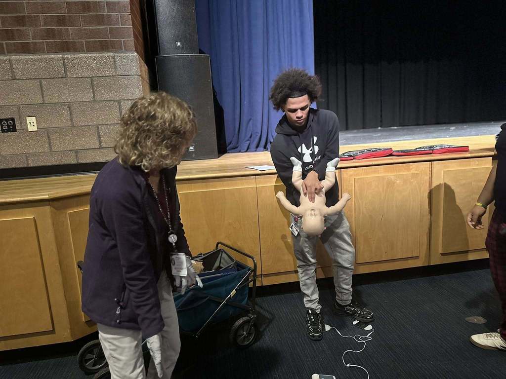 A student is practicing CPR on an infant mannequin in a school auditorium, while being guided by an instructor. 