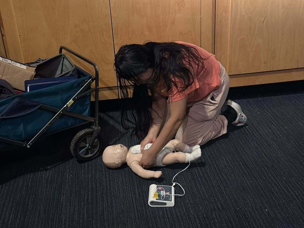 A student is keeling down on a dark carpeted floor in a school auditorium, practicing CPR on an infant mannequin.