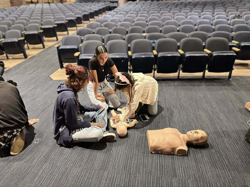 Three students are sitting down on a dark carpeted floor in a school auditorium, practicing CPR on an infant mannequin. Another mannequin can be seen nearby.
