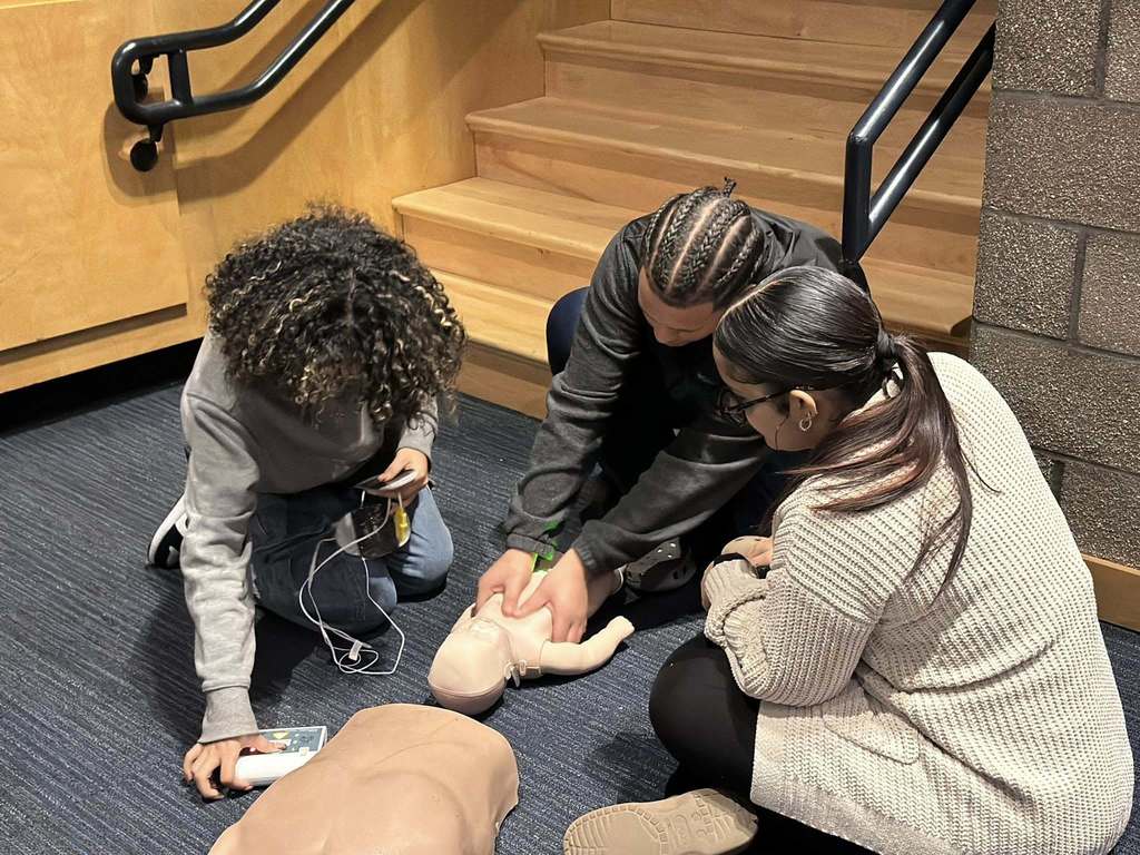 Three students are sitting down on a dark carpeted floor, practicing CPR on a baby mannequin in a school auditorium. One of the students is performing chest compressions while the other two students are observing.