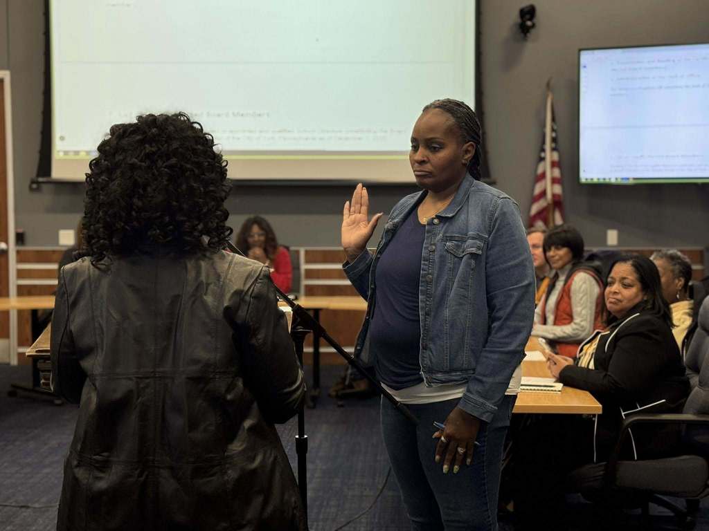 A woman is raising her right hand to take an oath in a conference room, facing an officiant. Other people can be seen in the background sitting at tables.