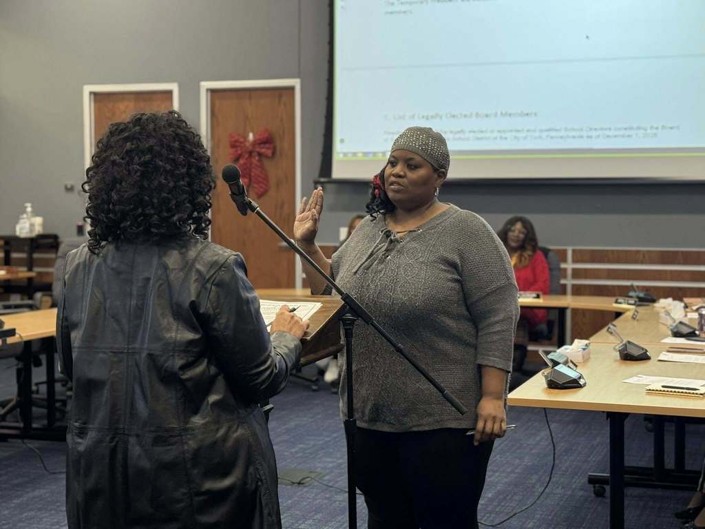 A woman is raising her right hand to take an oath in a conference room, facing an officiant. Other people can be seen in the background sitting at tables.