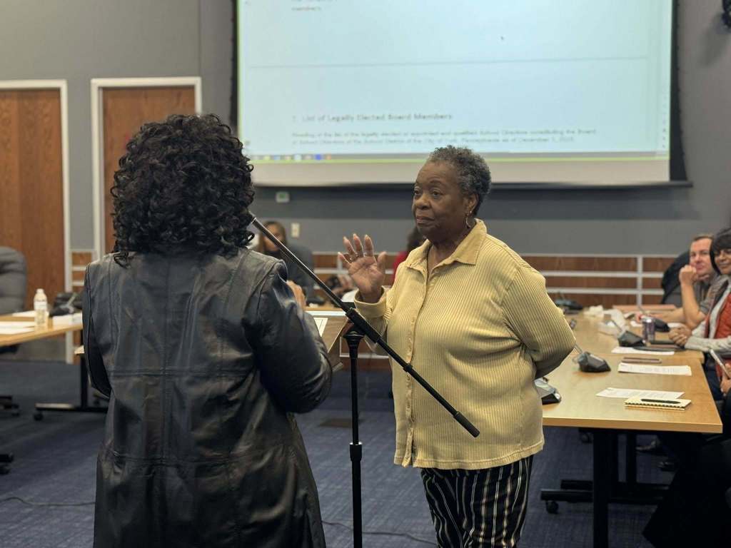 A woman is raising her right hand to take an oath in a conference room, facing an officiant. Other people can be seen in the background sitting at tables.