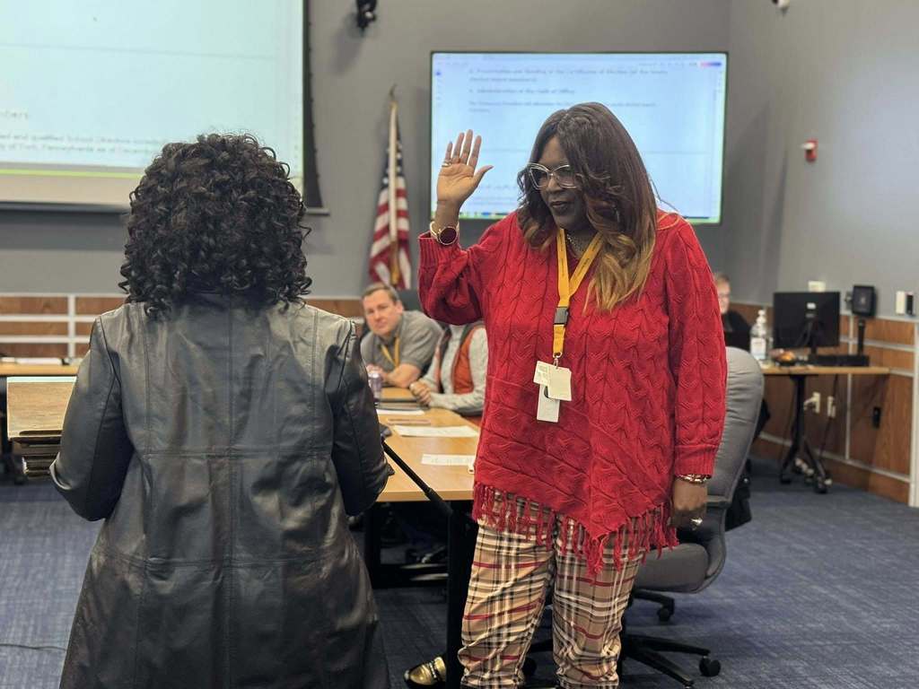 A woman is raising her right hand to take an oath in a conference room, facing an officiant. Other people can be seen in the background sitting at tables.