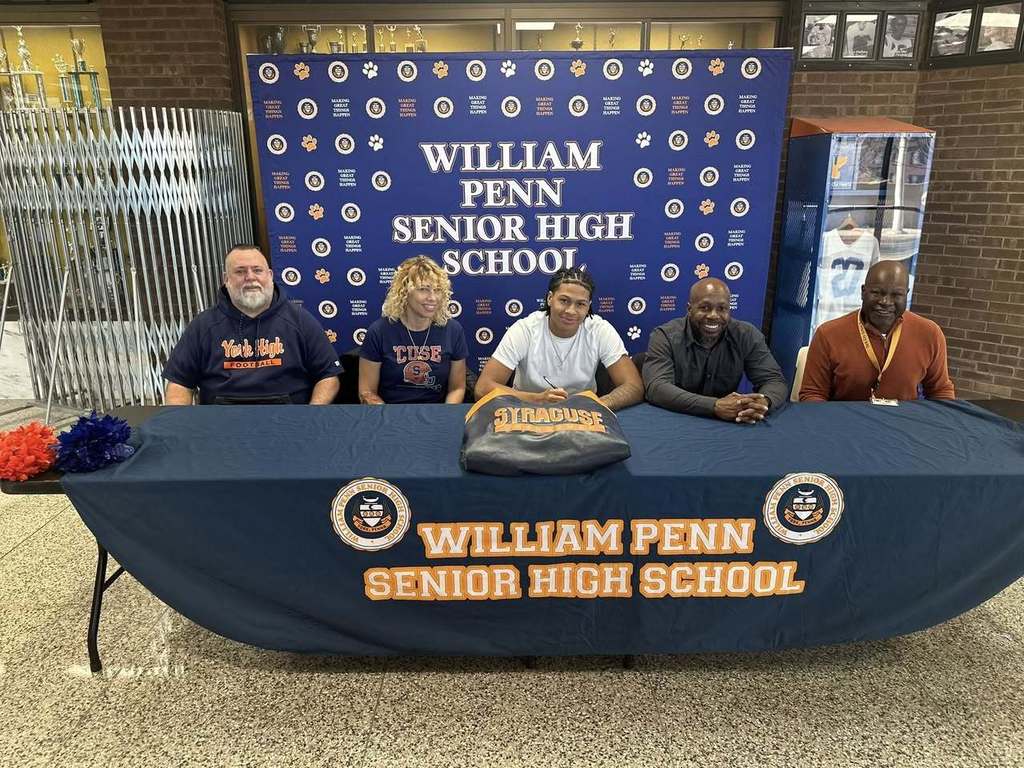 Five people, including district staff members, are sitting at a table with a "William Penn Senior High School" banner. A hoodie with "Syracuse" is on the table, suggesting a sports signing event.
