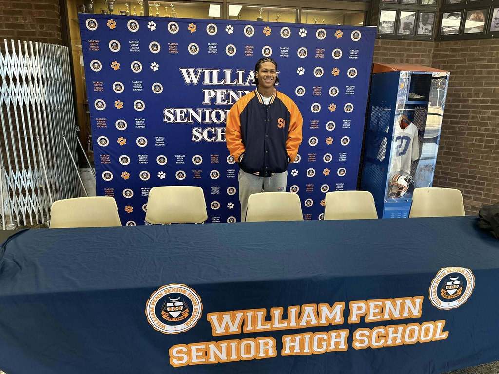 A student is standing in front of a William Penn Senior High School backdrop. He is wearing an orange and navy varsity jacket with the letters "S" and "U". A table with a matching banner is in front.