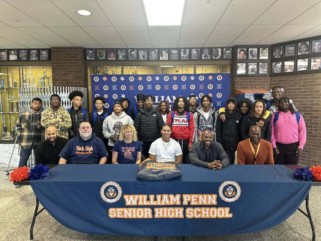 A group of students and adults, including district staff members are standing and sitting around a table at William Penn Senior High School. The table has a blue cloth with orange text and a "Syracuse" hoodie. Trophy cases and a photo wall are in the background.