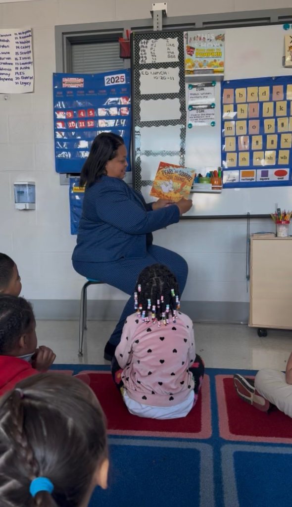 Dr. Andrea Berry-Brown, Superintendent of Schools, is sitting on a chair reading a book to a classroom of young students who are sitting on a colorful rug. 