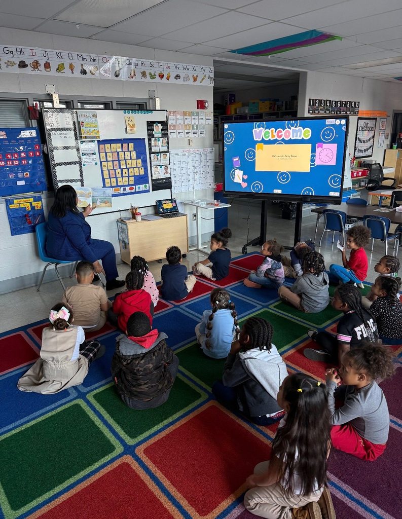 Dr. Andrea Berry-Brown, Superintendent of Schools, is sitting on a chair reading a book to a classroom of young students who are sitting on a colorful rug. A screen displays a "Welcome Dr. Berry-Brown" slide.