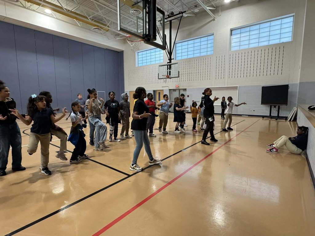 Two instructors are performing dance moves in front of a group of students who are observing them in a school gymnasium. A student can be seen sitting down nearby observing.