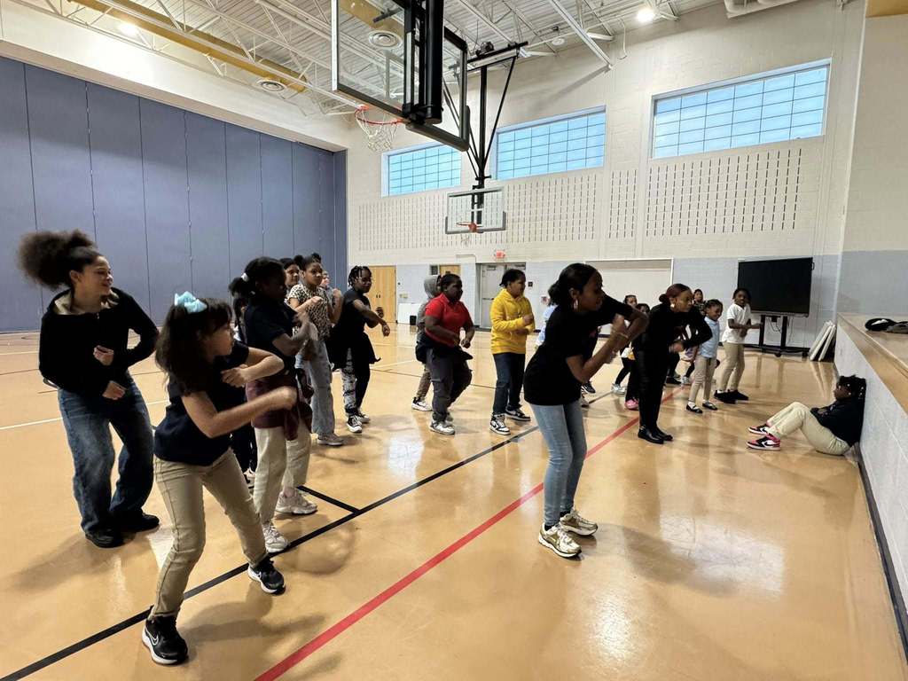 Two instructors are performing dance moves in front of a group of students who are observing them in a school gymnasium. A student can be seen sitting down nearby observing.