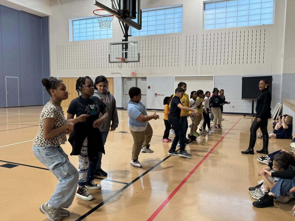 A group of students are performing dance moves in a school gymnasium while an instructor is observing them. A group of students can be seen sitting down nearby observing.