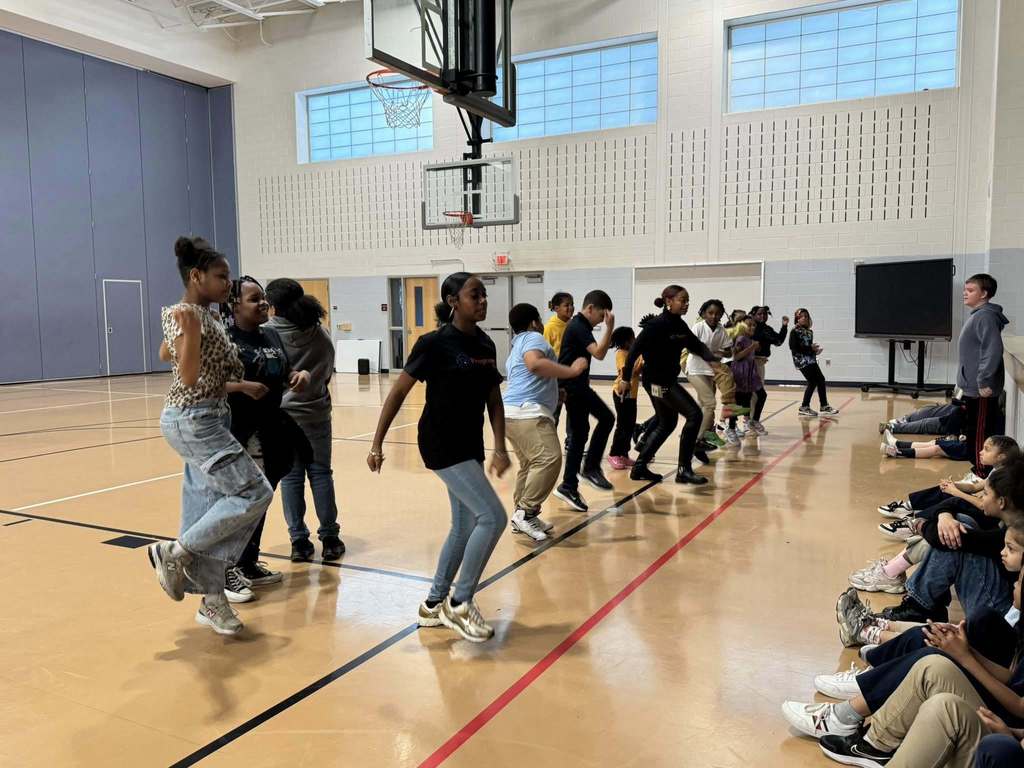 Two instructors are performing dance moves in front of a group of students who are observing them in a school gymnasium. A group of students can be seen sitting down nearby observing.