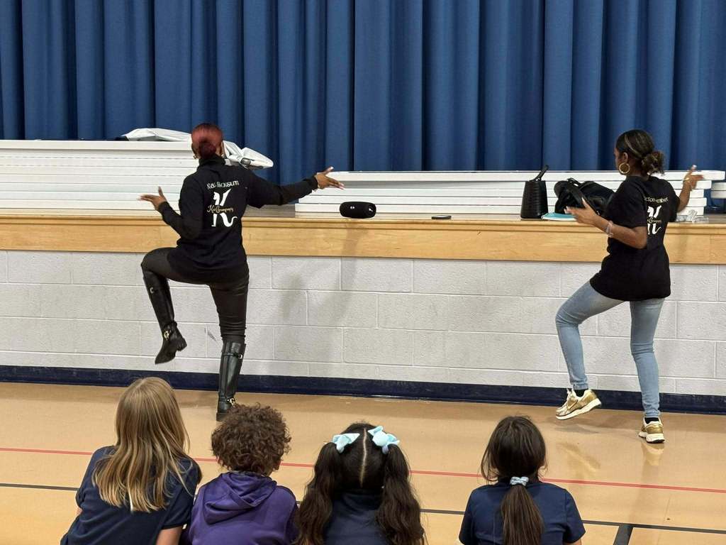 Two instructors are performing dance steps for a group of attentive students who are seated on the floor observing in a school gymnasium.  
