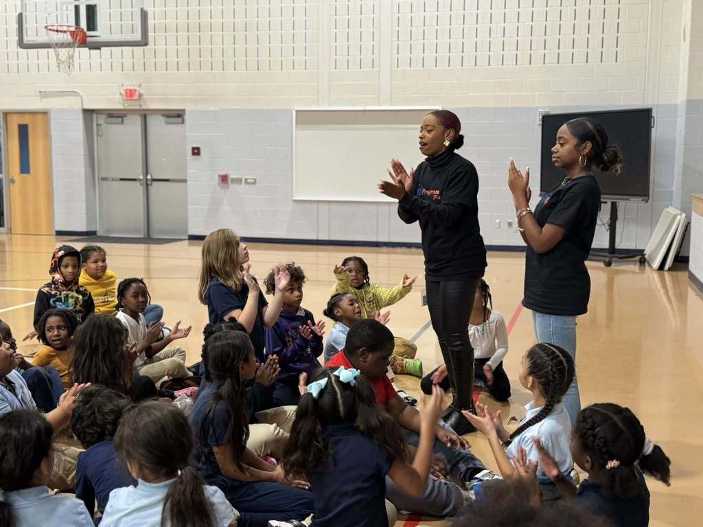 Two instructors are clapping and engaging with a group of attentive students who are seated on the floor in a school gymnasium. 