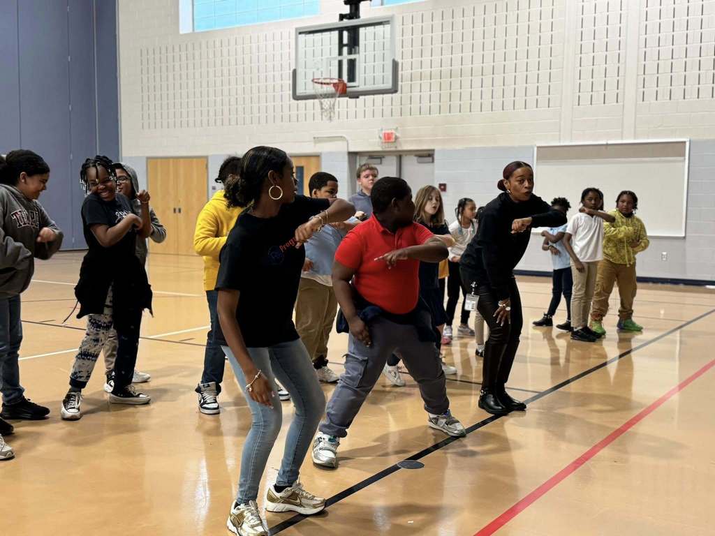 A group of students and two instructors are engaging in a dance class in a school gymnasium. They are moving in sync. A basketball hoop can be seen in the background.