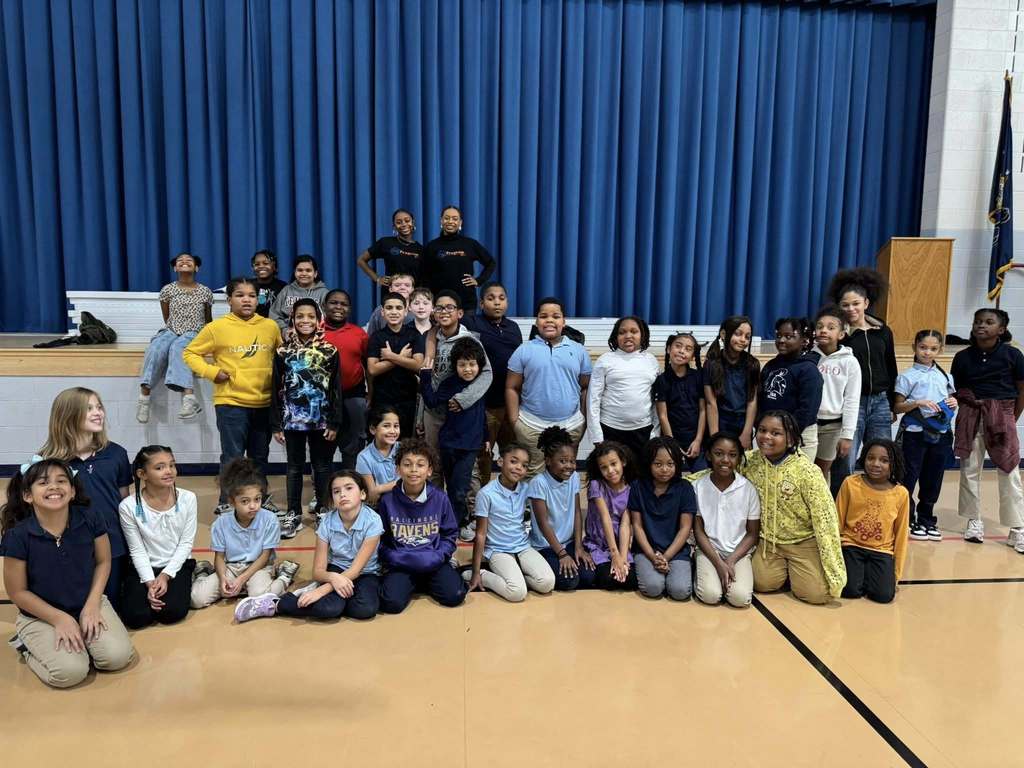 A group of students and two instructors are posing in a school gymnasium. A blue curtain backdrop can be seen in the background. 