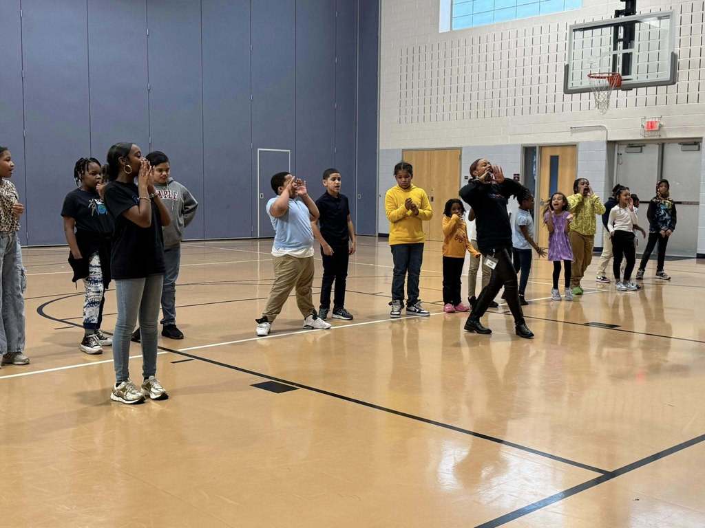 Two instructors are performing dance moves in front of a group of students who are observing them in a school gymnasium. 