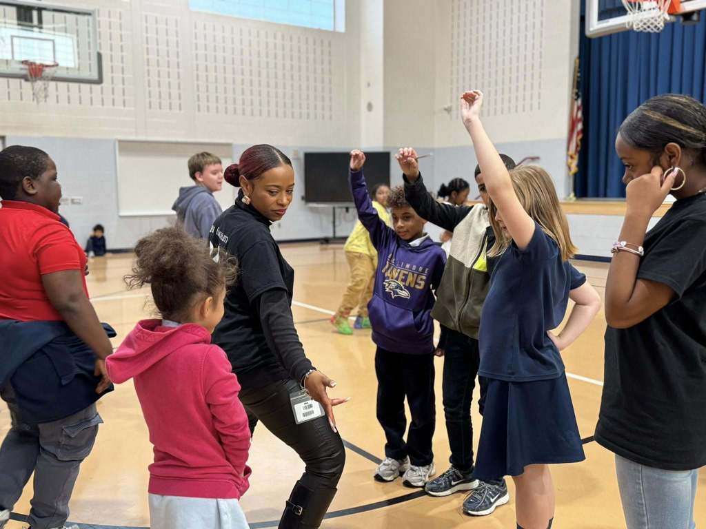 An instructor is leading a diverse group of students in a dance activity in a school gymnasium. The students are raising their hands enthusiastically.