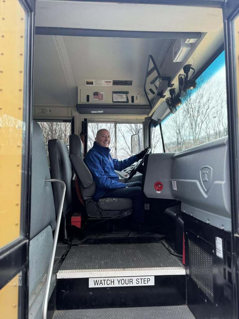 The School District of the City of York's Business Manager, Shawn Hain, is sitting in a yellow school bus driver's seat with one of his hands on the steering wheel. A "Watch Your Step" sign is visible.