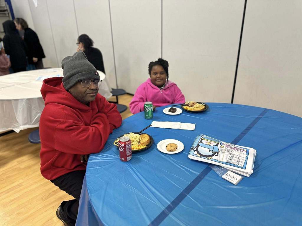 An adult and a young girl are sitting at a table that has plates of food, drinks and papers on it in a school cafeteria. Other people can be seen in the background.