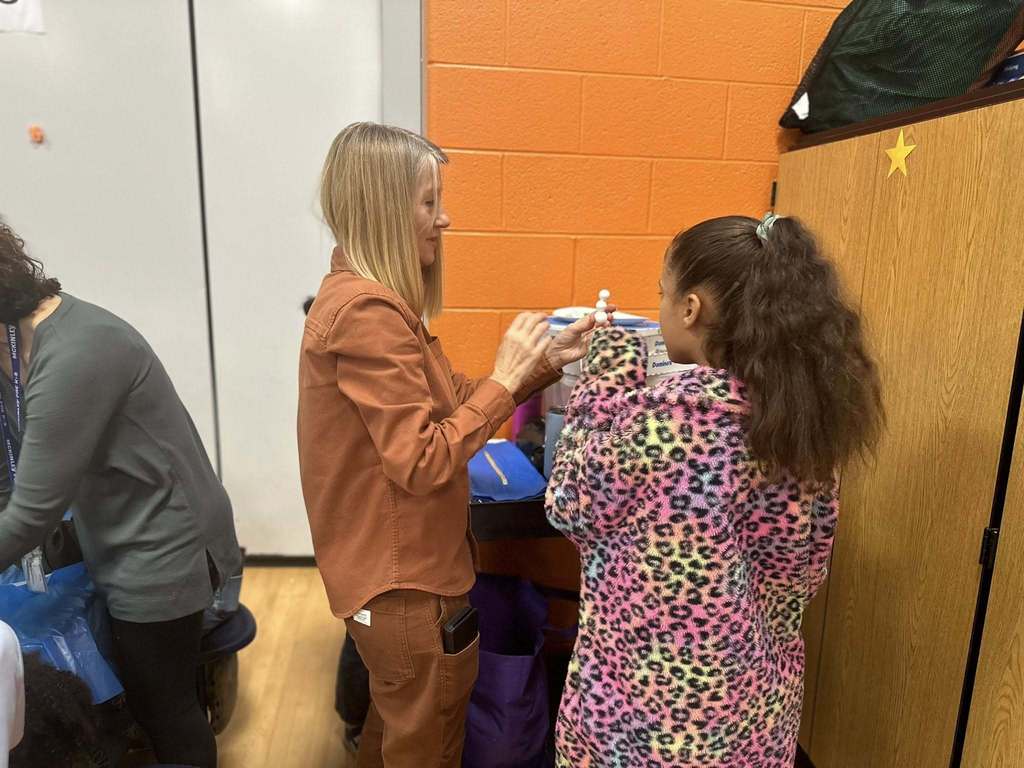 A district staff member is engaging in a craft with a young girl in a school cafeteria. Another district staff member can be seen nearby at a table.