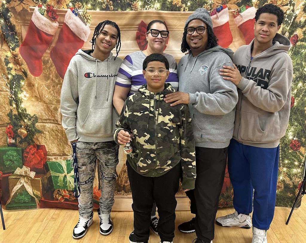A family of five, consisting of two adults, two teenagers, and one child, are standing in front of a festive backdrop that has stockings and presents on it. 