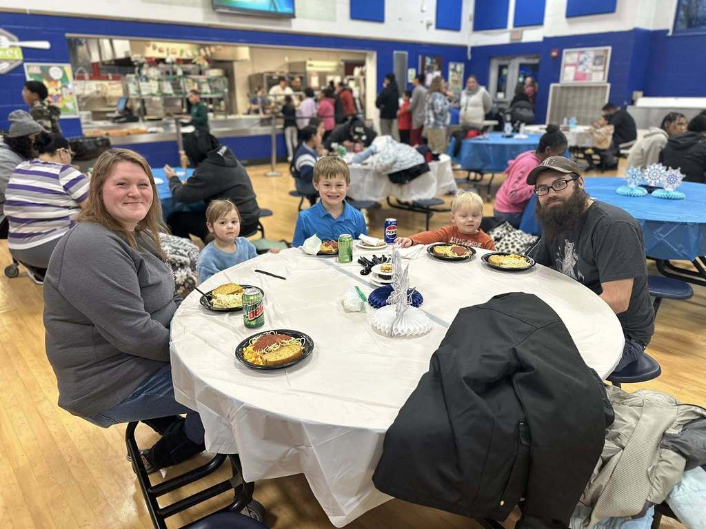 A family of five, consisting of two adults and three young children are sitting at a table with plates of food and drinks on it in a school cafeteria.