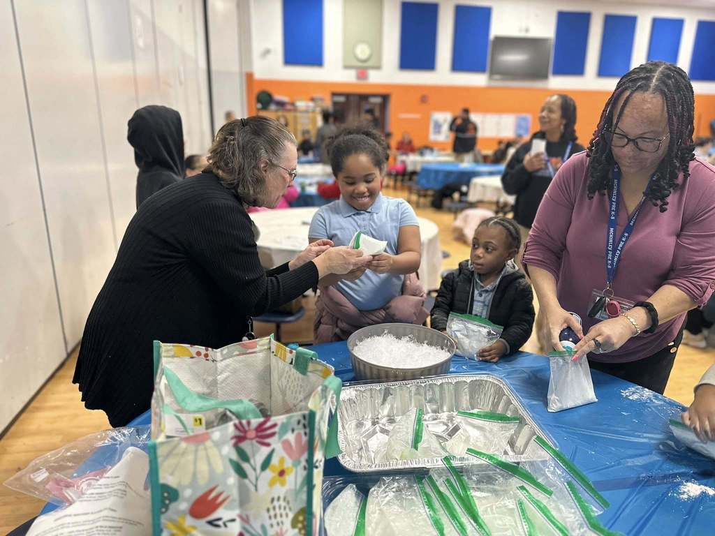 A group of two district staff members and two young children are gathered around a table in a school cafeteria making crafts. Other people can be seen in the background.