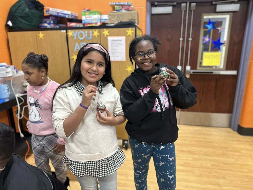 Two girls are holding up handmade ornaments in a school cafeteria. Another girl can be seen nearby in the background.