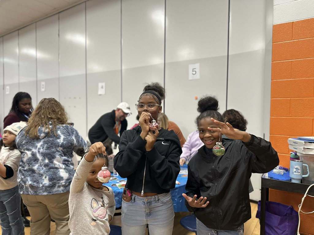 A group of three girls are holding up handmade ornaments in a school cafeteria. Other people can be seen behind them in the background.