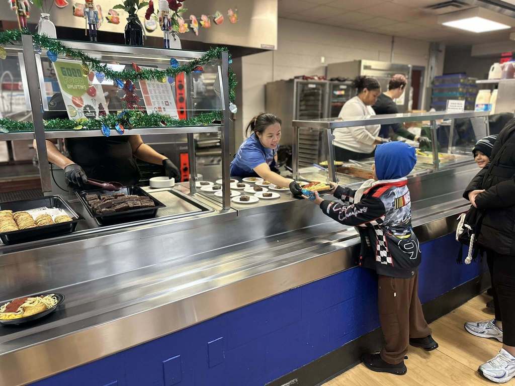 A district staff member is handing a plate of food to a young child across a counter in a school cafeteria. Other district staff members can be seen serving food in the background.