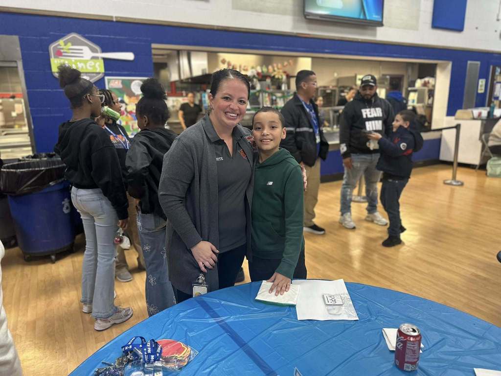 McKinley PreK-8 Principal, Dr. Ashley White, is standing next to a young boy behind a table in a school cafeteria. Other people can be seen in the background.