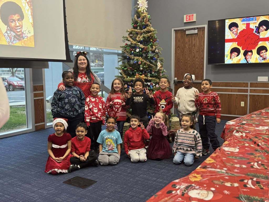 A district staff member and a group of children are gathered in front of a decorated Christmas tree, with holiday-themed images on screens nearby.