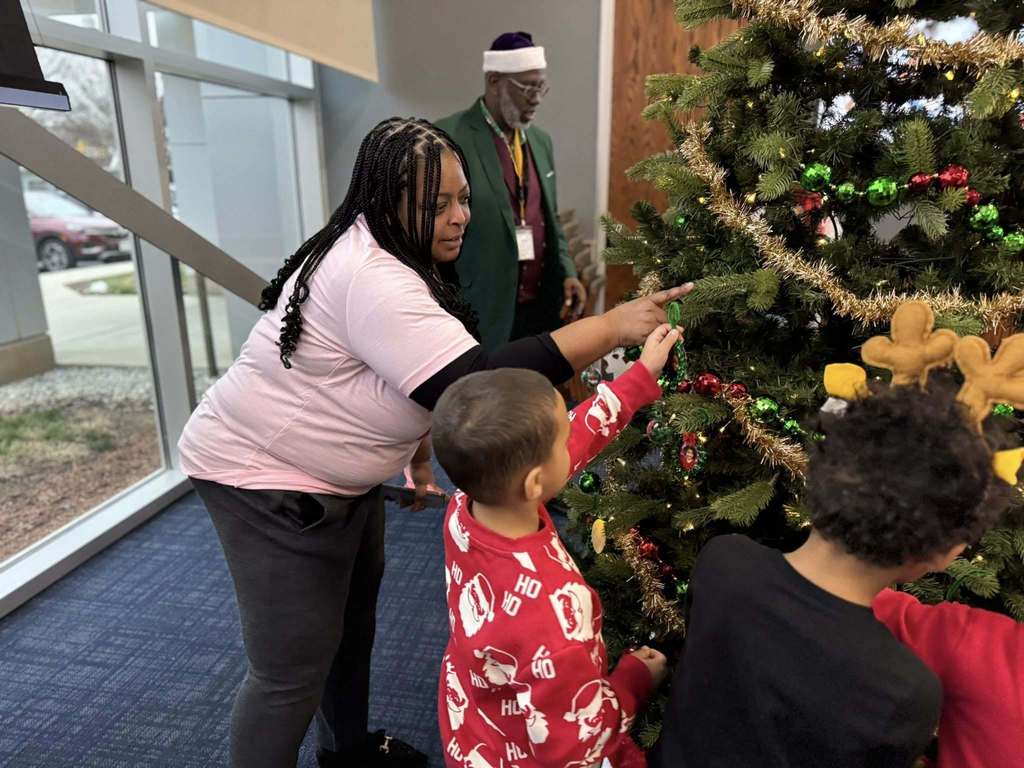 Dr. Andrea Berry-Brown, Superintendent of Schools, and tow young children are decorating a Christmas tree with colorful ornaments. Another district staff member can be seen in the background.