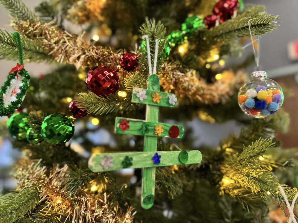A close-up of a decorated Christmas tree with colorful ornaments. A handmade green popsicle stick ornament is central, surrounded by red, green baubles, and a clear ornament filled with pom-poms. 