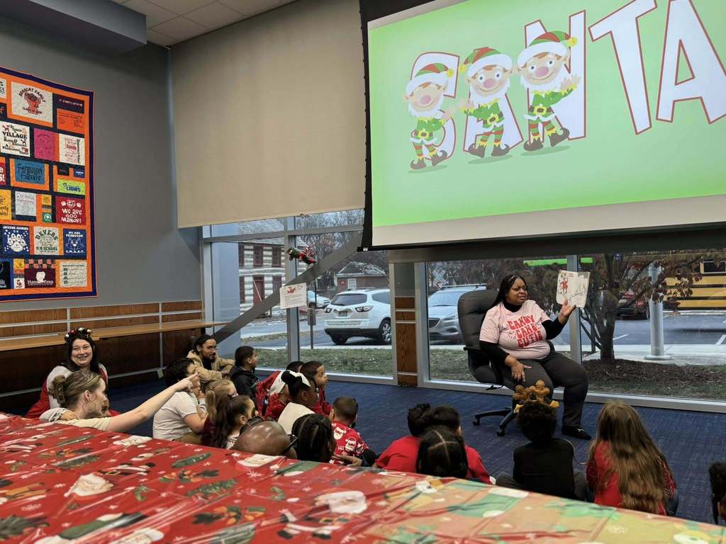 Dr. Andrea Berry-Brown, Superintendent of Schools, is reading a holiday book to an attentive group of students and district staff members. A large screen displays playful elves and the word "SANTA."