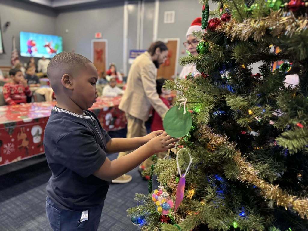A young boy is decorating a Christmas tree with ornaments in a festive room. Other people and children can be seen in the background.